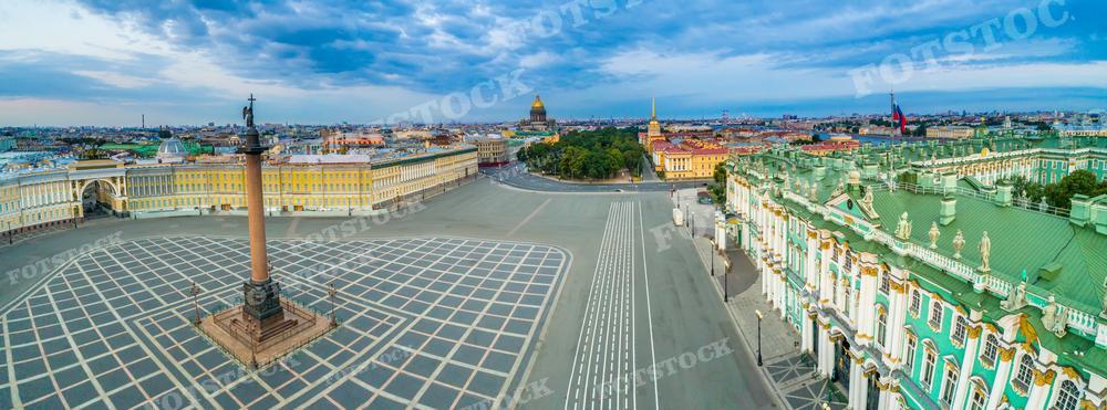 На панорамной фотографии представлена Дворцовая площадь в Санкт-Петербурге, взгляд на которую открывается с высоты птичьего полёта. В центре кадра находится Александровская колонна, окружённая геометрическими узорами площади. Слева виден знаменитый Зимний дворец, а справа — здание Главного штаба. На горизонте видны исторические силуэты городских достопримечательностей под облачным небом. Это фото может быть использовано для учебных материалов, туристических брошюр, в качестве изображений для статей о Санкт-Петербурге или как элемент городского пейзажа в дизайне интерьера.